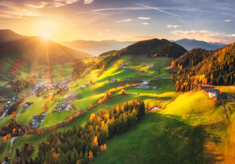 Fototapeta premium Santa Magdalena village valley with green fields in Dolomites, Italy at sunset. Aerial drone view of alpine autumn landscape with sunbeams, forest, houses, rolling hills and mountain range. Top view