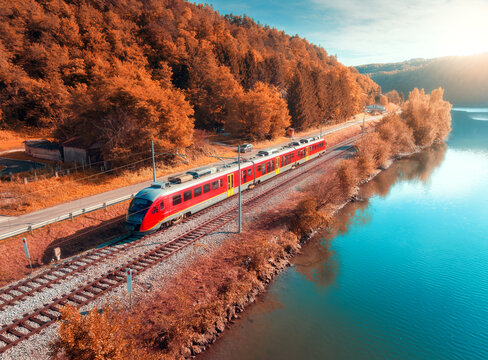 Aerial view of red passenger train riding on railway tracks and turquoise river in Slovenia. Top drone view of train, autumn forest, blue sky, clear water at golden sunset. Railroad, orange trees