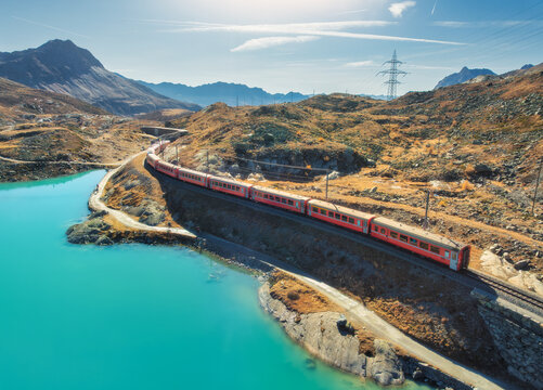 Aerial view of modern red train, turquoise mountain lake and blue sky at sunset in autumn. Bernina Express, Switzerland. Top drone view of train in Alps, winding railroad, orange hills in fall. Travel