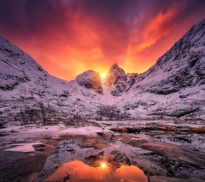Dramatic red sunset sky over snow covered mountains in Lofoten, Norway. Winter arctic landscape with sun flare behind peaks, vibrant orange clouds, rocks, ice pool and water reflection