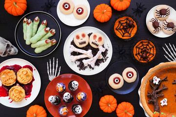 Fototapeta premium Fun Halloween dessert table scene over a black wood background. Top view. Selection of cookies, cupcakes and pumpkin and apple tarts and pies.