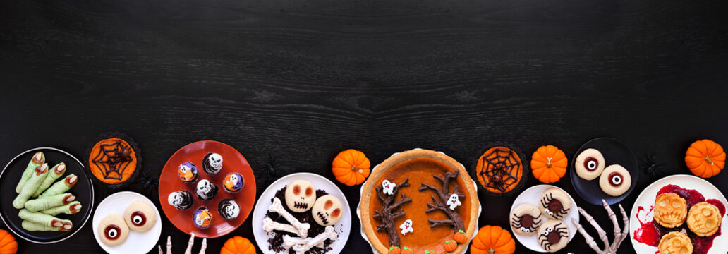 Fun Halloween dessert bottom border over a black wood banner background with copy space. Top view. Selection of cookies, cupcakes and pumpkin and apple tarts and pies.