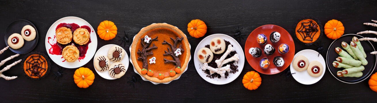 Fun Halloween dessert table scene over a black wood banner background. Above view.  Assortment of cookies, cupcakes and pumpkin and apple tarts and pies.