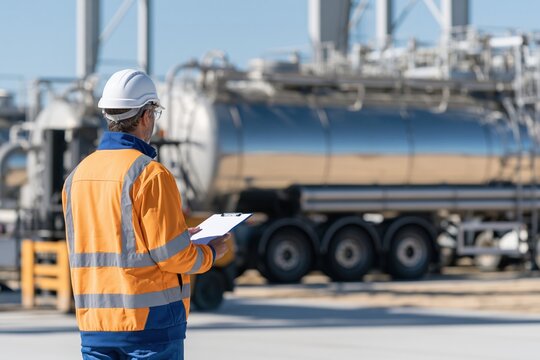 Worker in safety jacket and hard hat checking a clipboard with a large chemical tanker truck in a weathered industrial background for logistics and safety