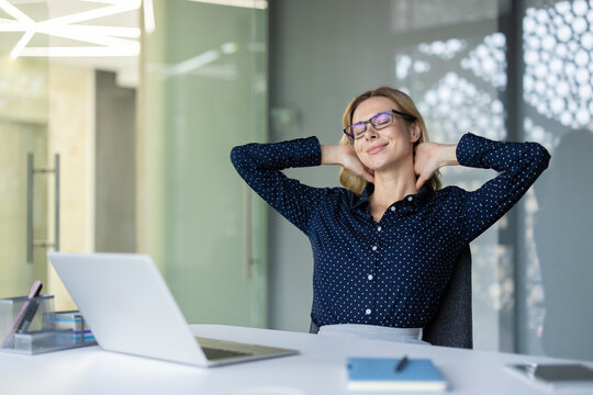 Businesswoman relaxing at her modern office desk, stretching arms behind head and smiling with closed eyes, finding relief from stress during a well-deserved work break