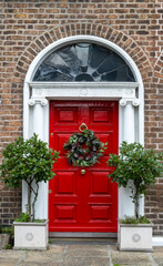 Obraz premium Red Georgian front door decorated for Christmas Holiday season with seasonal wreath. Entrance to house with decorative fanlight, columns and round bay laurel trees. Dublin, Ireland
