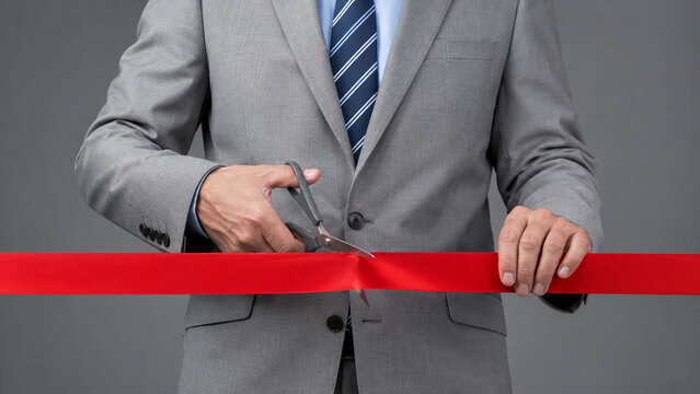 A businessman in a grey suit cuts a bright red ribbon with scissors during an inauguration ceremony against a grey background.