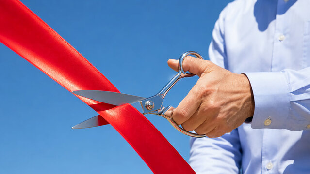A person in a light blue shirt uses large silver scissors to cut a bright red ribbon against a clear blue sky.