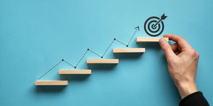 Hand arranging wooden block stairs with rising graph line leading to target icon. Business growth, step-by-step strategy, and goal achievement concept.