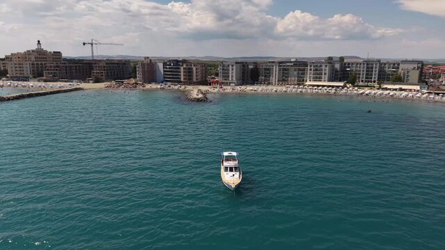 Scenic high-angle aerial 4K drone video of a white leisure boat yacht cruising in the blue sea water near the sandy beach, hotels, and apartments of Nesebar coastal resort in Bulgaria on a clear sunny