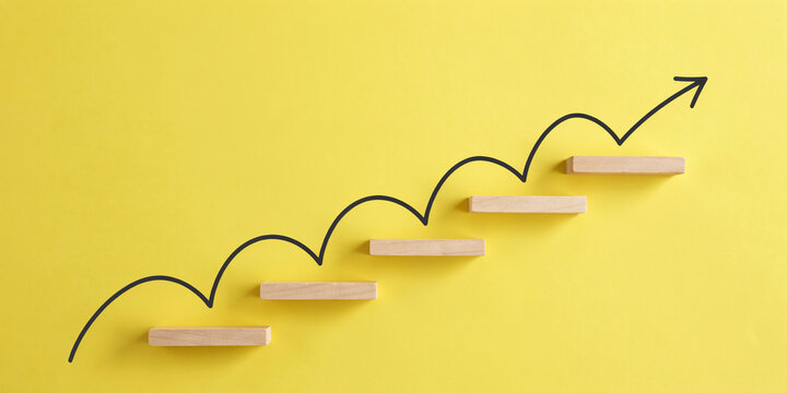 Wooden block stairs with curved upward arrow line on yellow background. Step-by-step business growth, market development, and progress concept.