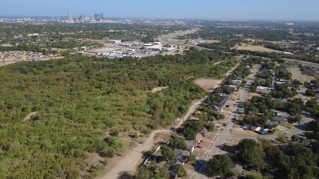 Expansive green parkland borders sunny neighborhood street grids across Historic Southside. Dense tree lines stretch toward distant downtown city skyscrapers under sun, Fort Worth, Texas