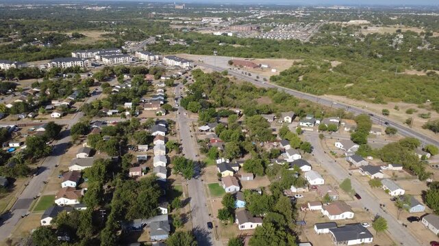 Paved neighborhood thoroughfares link quiet residential homes across DFW Metroplex. Clustered rooftops blend into dense backyard foliage under open midday daylight, Fort Worth, Texas