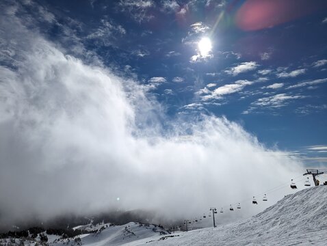 Towering mountain fog rolls across a sunlit ski slope beneath brilliant blue skies as chairlifts climb into the advancing cloud bank during a dramatic winter day