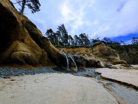 A small freshwater cascade spills down sculpted sandstone cliffs onto a broad sandy beach along the Oregon Coast, beneath windswept evergreen trees and dramatic low clouds