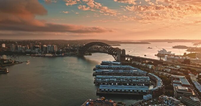 Amazing colorful sunset over Sydney cityscape showcasing the iconic Harbour Bridge and Opera House, with boats navigating the harbor waters under a warm, glowing sky. Aerial view drone flight footage