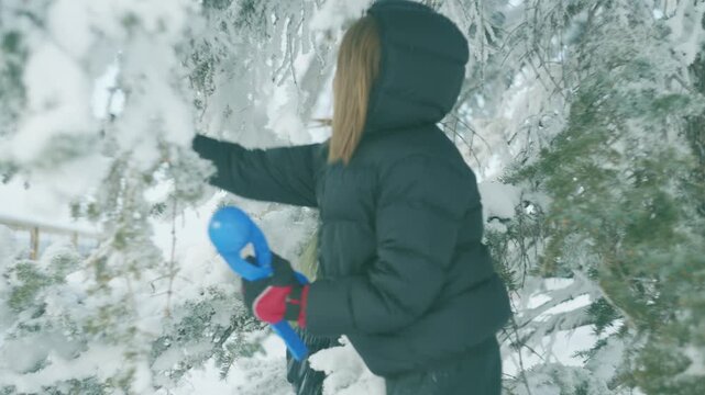 Boy in a puffer jacket carrying a blue snowball maker while walking among snowy pine trees