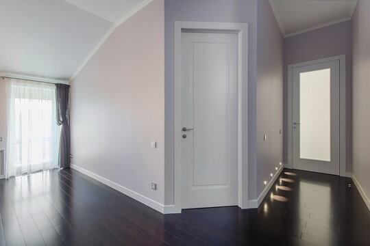 A hallway inside a modern building features light-colored walls and dark wooden floors. Natural light comes in from windows on one side. Doors are present on both walls
