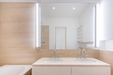 Fototapeta premium Two sinks are set on a simple countertop with smooth edges. Bright lights illuminate the space. A large mirror reflects the room. The decor is minimal and practical