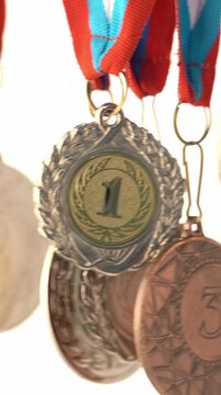 First-place medal with a "1" is central. Silver and bronze medals hang nearby. Ribbons are red and blue. Shiny metal surfaces glow. Background is softly blurred