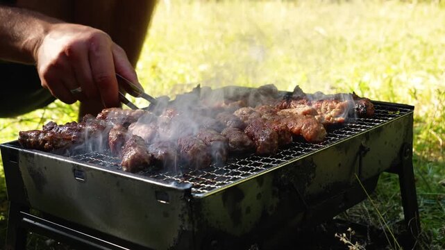A man turns cevapi with tongs on a smoky charcoal grill outdoors. Traditional barbecue footage suits food, travel, leisure and hospitality themes