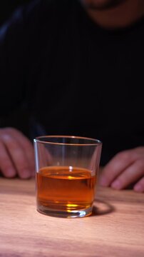 Close-up of amber liquid in a clear glass on wood table. Person's hands rest nearby, suggesting quiet contemplation. Ideal for scenes of relaxation, solo drinking, or quiet moments