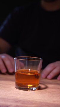 Close-up of amber liquid in a clear glass. Hands rest gently on a wooden table. Calm posture suggests quiet reflection. Ideal for scenes of relaxation or social moments. Focus on the drink's texture