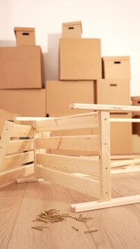 Light wood chair frame lies on wooden floor. Scattered screws surround the frame. Cardboard boxes stack in the background. Furniture assembly work is underway