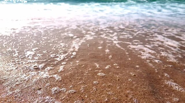 Close-up low-angle shot of ocean water washing over golden sand, leaving shimmering bubbles and white foam under bright daylight.