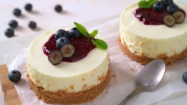 Two mini cheesecakes rest on parchment paper, adorned with vibrant berry sauce, fresh blueberries, and mint leaves, illuminated by soft natural lighting.