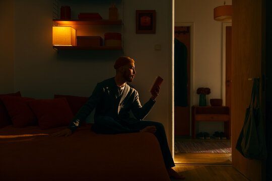 Young man in beanie sitting on bed reading book in cozy dimly lit warm apartment room at evening enjoying peaceful solitary literature study relaxation night hobby inside domestic ambient dark home