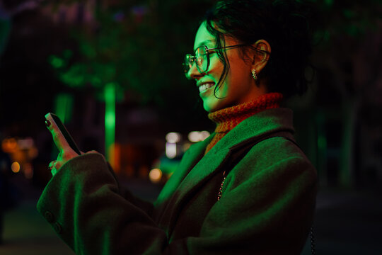 Cheerful multiracial woman texting on smartphone in urban park at night, green light background, mobile application user, authentic ethnic representation of young professional, side view.