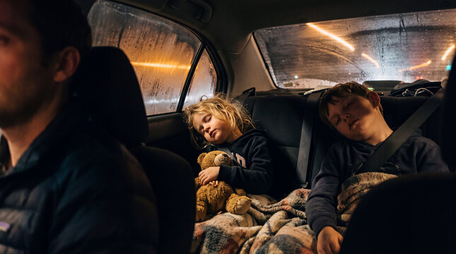 Two children are sleeping in the back seat of a car. An adult is driving in the front. The car is moving at night with light shining through the windows