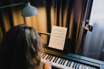 Woman playing digital piano with sheet music during an online video lesson on a smartphone, learning and practicing music remotely from home © VK Studio