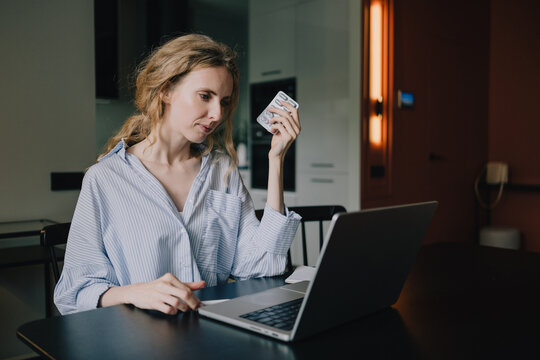 Woman holding a blister pack of pills while using a laptop for an online healthcare consultation at home