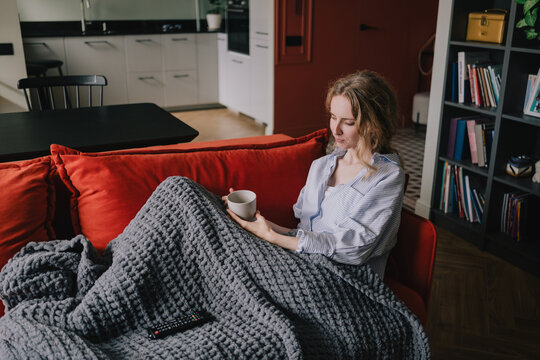Woman resting under a warm blanket with a cup while relaxing on a sofa at home