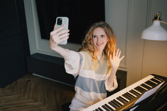 Smiling woman waving during a video call beside a digital piano at home, using smartphone technology for remote music learning and piano education