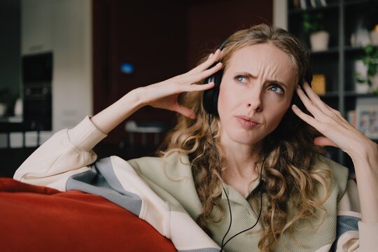 Woman wearing headphones at home with a disturbed expression, reacting to unwanted noise and seeking quiet while trying to block distracting sounds