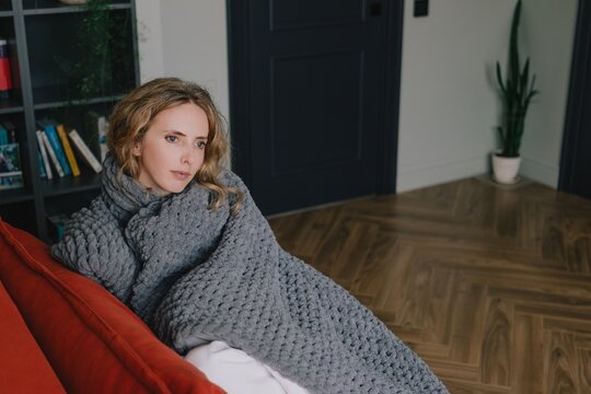 Woman wrapped in a thick blanket to keep warm while sitting on a sofa at home