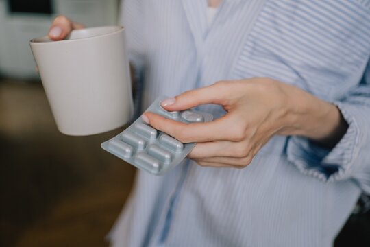 Close-up of woman holding a blister pack of pills and a cup at home, preparing to take medication as part of an everyday personal healthcare routine