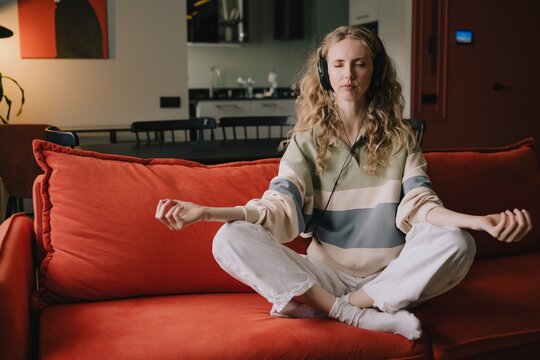 Woman meditating with headphones while sitting cross-legged on a sofa at home, practicing mindfulness, relaxation and self-care for emotional wellbeing