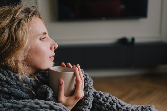 Woman wrapped in a thick blanket holding a cup to keep warm while resting at home