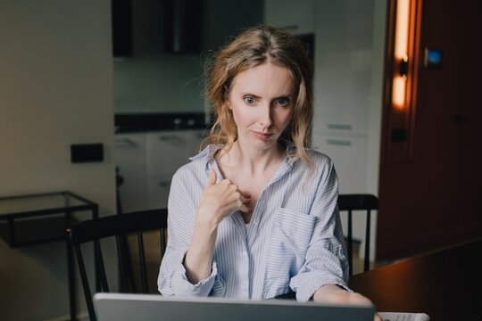 Woman pointing to herself with a questioning expression during an online conversation on laptop at home