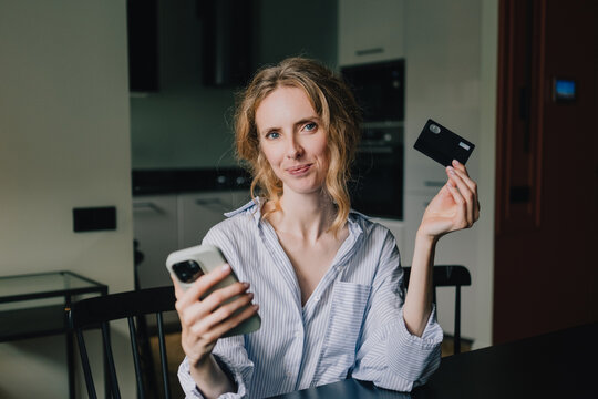 Woman holding smartphone and bank card after completing a successful online payment at home
