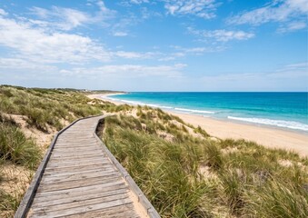 Obraz premium Wooden boardwalk leading through grassy sand dunes to a scenic tropical beach and blue ocean