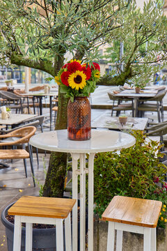 Beautiful autumn bouquet with sunflowers and red dahlias in brown glass vase on white high street cafe table with wooden bar stools and olive tree in background on a rainy day in Germany.