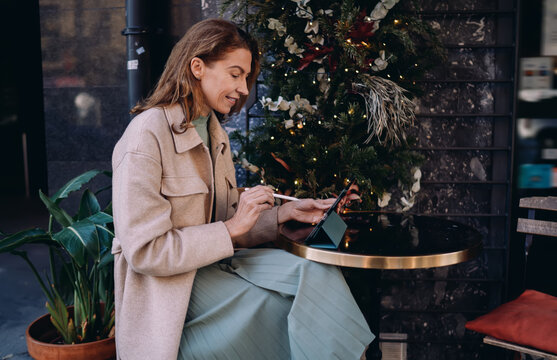 Mature woman using digital tablet with stylus sitting at cafe table near decorated Christmas tree. Successful female architect or designer working remotely during holidays. Career and tech.