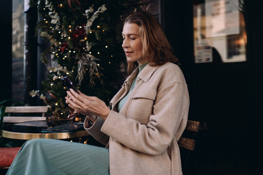 Profile of mature woman using smartphone in street cafe, Caucasian freelancer typing message on mobile phone, digital communication and remote work concept, stylish lady in autumn setting.