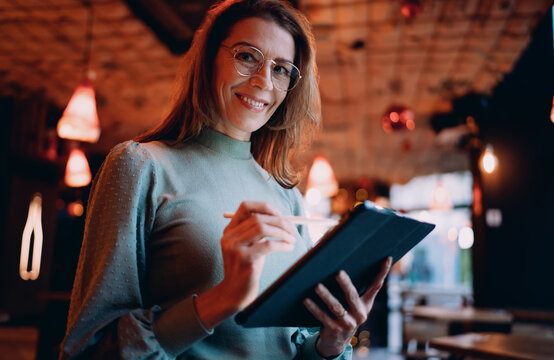 Smiling Caucasian woman looking at camera with tablet and stylus, professional freelancer working at cafe, career growth and digital nomad lifestyle, authentic representation, real people.