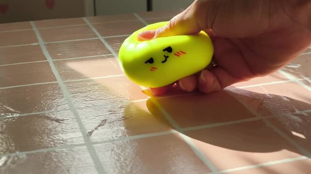 Hand gently squeezes a bright yellow squishy toy resembling a dumpling on a glossy tiled surface, showcasing the playful deformation and light reflections in a cozy indoor setting